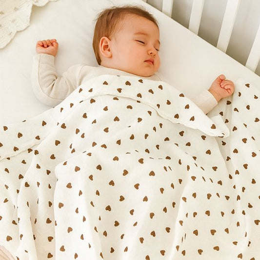 Bébé endormi paisiblement sous une couverture blanche à motifs de petits cœurs marron – douceur et confort garanti.
