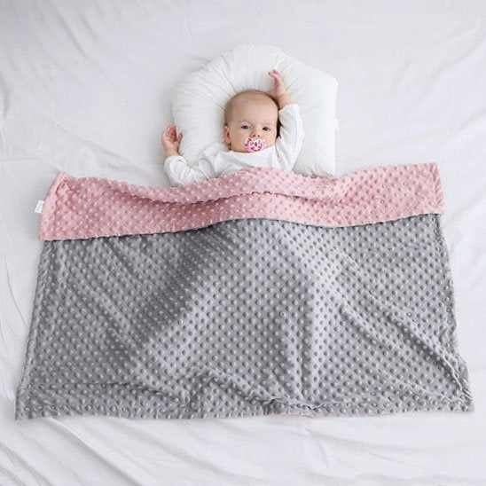 Bébé allongé paisiblement sous une couverture double face rose et grise, parfaite pour le sommeil et l’emmaillotage
