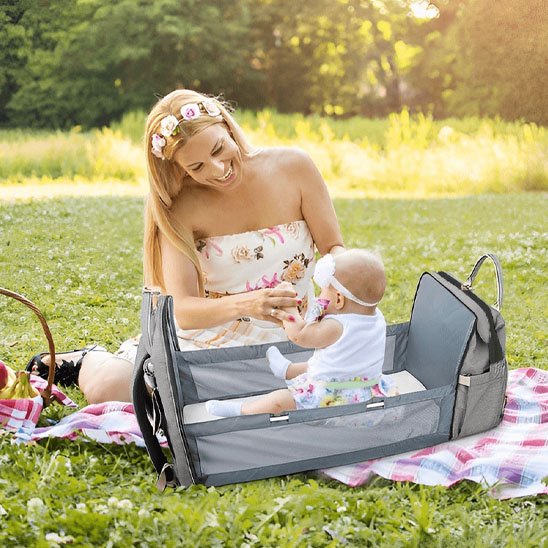  Maman souriante assise sur une couverture en plein air jouant avec son bébé dans un sac à langer convertible en lit portable, ambiance pique-nique.