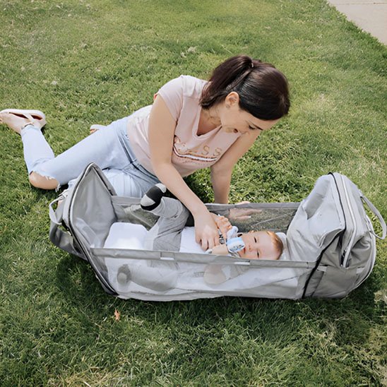  Maman changeant son bébé dans un lit portable gris intégré au sac à langer, moment de soin en plein air.